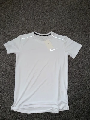 Vit Nike löpartröja herr - Snygg vit löpartröja från Nike med korta ärmar och reflekterande detaljer på axlarna. Tillverkad i ett lätt och snabbtorkande material som passar perfekt för träning. Nike-logga på bröstet och 'Nike Running' tryckt i nacken.