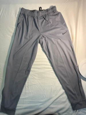 Gråa mjukisbyxor från Nike - I stort sätt oanvända. Enbart testade. Snygga grå mjukisbyxor från Nike med smal passform och dragkedja vid benslut. Byxorna har resår i midjan och är tillverkade i ett mjukt, stretchigt material som känns skönt mot huden. Perfekta för chill eller träning. Diskret Nike-logga på låret. 