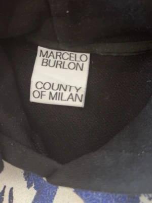 Marcelo Burlon svart hoodie med vingar - Svart hoodie från Marcelo Burlon County of Milan med ikoniskt vingtryck i vitt och blått över axlar och bröst. Huva med dragsko och klassisk passform. Perfekt för dig som vill sticka ut med streetwear-vibe.