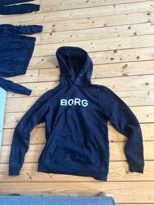Marinblå hoodie från Björn Borg - Snygg marinblå hoodie från Björn Borg med stor vit BORG-logga på bröstet. Klassisk känguruficka framtill och justerbar huva med dragsko. Tillverkad i mjuk bomullsblandning som är skön att ha på sig. Perfekt för chill dagar eller när du vill ha en clean look.