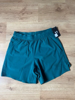 Under Armour shorts - Snygga gröna träningsshorts från Under Armour med elastisk midja och diskret logga på linningen. Shortsen har en loose passform och är tillverkade i ett lätt och snabbtorkande syntetmaterial, perfekta för sommaren. Köpt nya för 749kr, säljer pga för liten storlek. Helt nya och oanvända.