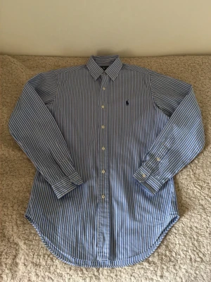 Ralph Lauren Skjorta - Klassisk blårandig skjorta från Ralph Lauren med button-down krage och långa ärmar. Skjortan har vita knappar och den ikoniska broderade loggan på bröstet. Tillverkad i bomull för en skön och luftig känsla. Perfekt till jeans eller chinos.