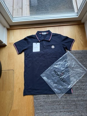 Marinblå pikétröja från Moncler - Snygg marinblå pikétröja från Moncler med klassisk krage och knappar framtill. Tröjan har vita och röda detaljer på krage och ärmslut samt Moncler-logga på bröstet. Tillverkad i bomull för en bekväm känsla.