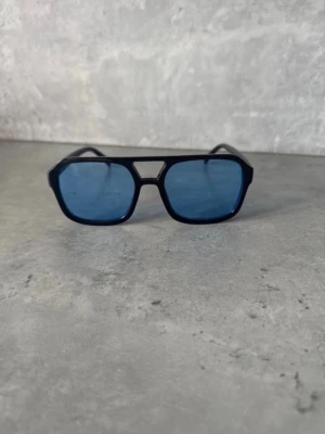 Svarta solglasögon med blått glas - Snygga solglasögon med svart, kantig båge och blåtonade glas. Modellen har en modern, geometrisk form och bred brygga. Passar perfekt för dig som vill sticka ut med en cool accessoar. Tillverkade i plast för en lätt och bekväm känsla.