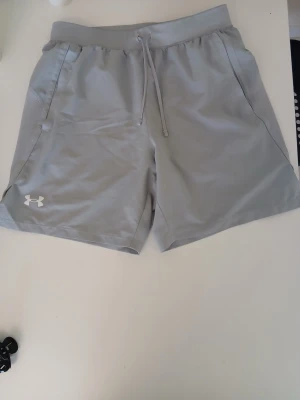 Ljusgrå shorts från Under Armour - Snygga ljusgrå shorts från Under Armour med elastisk midja och snörning. Tillverkade i ett lätt och snabbtorkande syntetmaterial, perfekta för träning eller chill. Klassisk logga på vänster ben och praktiska sidofickor.