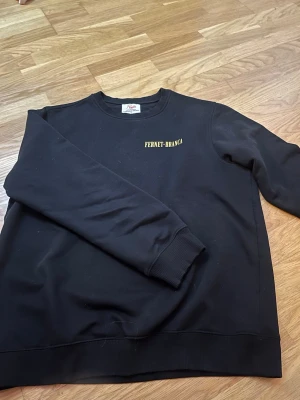 Svart Fernet-Branca sweatshirt - Cool svart sweatshirt med gult Fernet-Branca-tryck på bröstet och stort tryck på ryggen med logga och text. Tröjan har rund hals, långa ärmar och ribbade muddar. Perfekt för dig som gillar streetwear och vill sticka ut med en unik design.