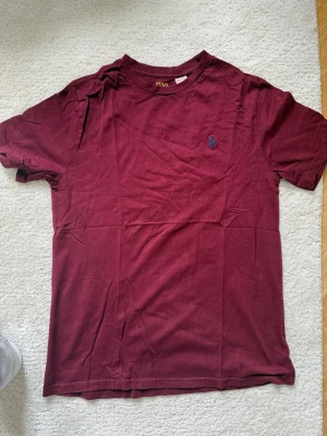 Vinröd t-shirt från Polo Ralph Lauren - Säljer en vinröd t-shirt från Polo Ralph Lauren med klassisk passform och liten blå logga broderad på bröstet. T-shirten har rund halsringning och är tillverkad i mjuk bomull, perfekt för en clean och stilren look.