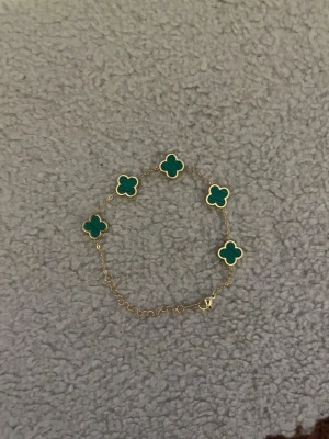 Guldarmband med gröna klöverhängen - Snyggt armband i guldton med fem gröna fyrklöverformade hängen. Kedjan är tunn och smidig med ett klassiskt lås. Perfekt accessoar för dig som gillar stilrena detaljer och färgklickar i din outfit.