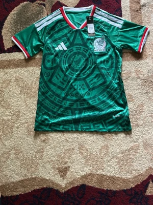 Fotbollströja  - Snygg grön Mexiko landslagströja från Adidas med Aztek-inspirerat mönster, vita axelränder och röd-vit krage. Tröjan har Mexikos fotbollsförbunds logga på bröstet och är tillverkad i lätt, ventilerande material (AEROREADY). Perfekt för match eller träning.