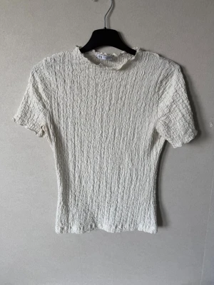 Vit ribbad topp från Zara - Säljer en vit ribbad topp från Zara med korta ärmar och hög hals. Toppen har en stretchig struktur och är tillverkad i ett mjukt material som sitter snyggt på kroppen. Perfekt att styla med jeans eller kjol.
