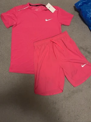 Rosa träningsset från Nike - Säljer ett sportigt rosa träningsset från Nike med t-shirt och shorts. Båda plaggen har Nike-loggan tryckt i vitt och är tillverkade i ett lätt och snabbtorkande material. Perfekt för gymmet eller löprundan. Skriv för mer info