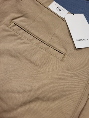 Beige chinos från These Glory Days 56 Xl - Snygga beige chinos från These Glory Days i klassisk rak modell. Byxorna har bälteshällor, två sidofickor och två bakfickor med minimalistisk design. Tillverkade i ett mjukt bomullstyg som ger en clean och stilren look.