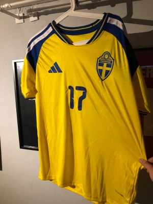 Svensk landslagströja Adidas gul - Säljer en gul svensk landslagströja från Adidas med blå detaljer och nummer 17 på framsidan. Tröjan har korta ärmar, rund hals och Sveriges emblem på bröstet. Materialet är lätt och andas, perfekt för fotboll eller träning.