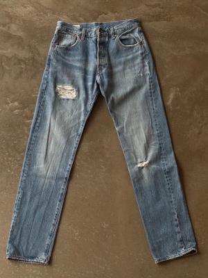Levi’s 501 ’93 Straight Jeans – Ljusblå Distressed (W30/L34 - Levi's 501 i '93-modell, vilket innebär något högre midja och helt raka ben. Ljusblå tvätt med fabriksgjort slitage/hål över knän och lår. Traditionell knappgylf och läderpatch baktill.