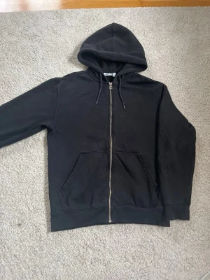 Svart zip-hoodie från Weekday - Svart hoodie från Weekday med dragkedja framtill, huva med dragsko och två stora fickor. Klassisk och enkel design i mjukt material, perfekt för en avslappnad stil.