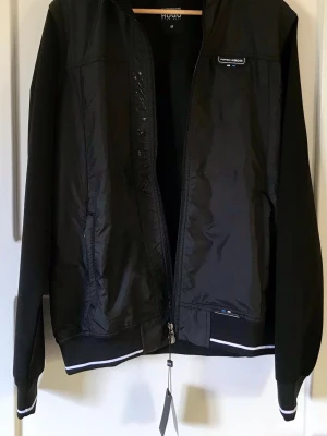 Svart vindjacka Porsche x Hugo Boss med byxor - Snygg svart vindjacka från Porsche x Hugo Boss med dragkedja framtill och diskreta loggor på bröstet och insidan. Jackan har ribbade muddar med vita detaljer och är tillverkad i ett lätt, vindtätt material. Perfekt för en sportig och stilren look. Byxor medföljer.