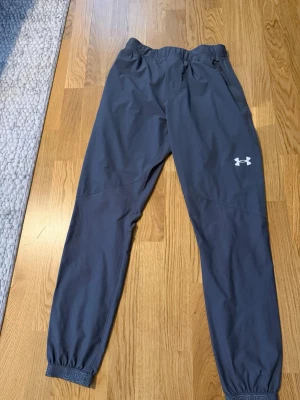 Mörkgrå träningsbyxor Under Armour - Snygga mörkgrå träningsbyxor från Under Armour med resår i midjan och benslut. Byxorna har en diskret logga på låret och är tillverkade i ett lätt och snabbtorkande material, perfekta för löpning eller gym. Smal passform och sportig look.
