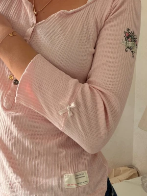 Unik Odd Molly topp - Säljer denna sällsynta odd Molly topp. Supersöt ljusrosa ribbad topp från Odd Molly med knappar framtill, liten rosett på ärmen och broderad detalj på armen. Toppen har lång ärm och en feminin, något v-ringad halsringning med spetskant. Perfekt för dig som gillar gulliga detaljer.