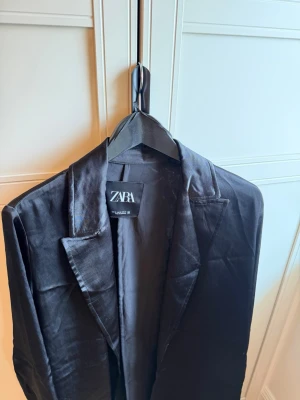 Svart kavaj från Zara - Stilren svart kavaj från Zara med klassisk krage och smal passform. Tillverkad i ett glansigt viskos material som ger en modern look. Perfekt för dig som vill ha en enkel men snygg kavaj till garderoben.