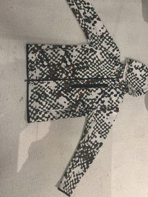 Nike camo teck fleece med huva - Nike vindjacka med hel dragkedja och huva i ett fett camomönster i vitt, svart, grönt och brunt. Långärmad modell med lätt material och sportig vibe. Perfekt för dig som vill ha en unik och trendig jacka till din streetwear-look.