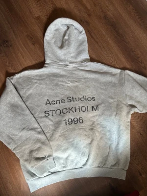 Grå hoodie Acne Studios Stockholm - Snygg grå hoodie från Acne Studios med tryck på ryggen där det står 'Acne Studios STOCKHOLM 1996'. Klassisk passform med huva och ribbade muddar. Tillverkad i mjuk bomullsblandning för maximal komfort. Perfekt för en avslappnad och trendig look.