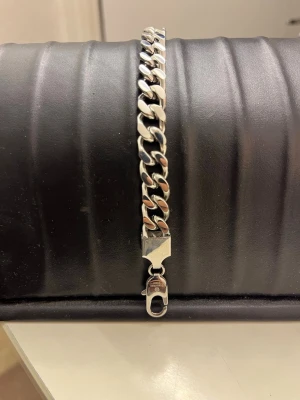 Silverfärgat kedjearmband i stål - Snyggt och stilrent armband i silverfärgat stål med breda, platta länkar och en robust låsdetalj. Perfekt accessoar för dig som gillar en clean och modern look. Passar både till klocka eller för sig själv.
