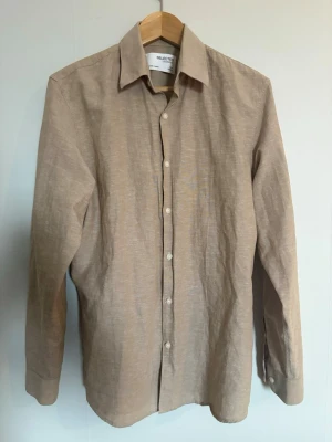 Beige linneskjorta från Selected Homme - Stilren beige linneskjorta från Selected Homme i svalt linnematerial. Klassisk krage, långa ärmar och knäppning framtill med vita knappar. Perfekt för en avslappnad men ändå uppklädd look. Varan är aldrig använd