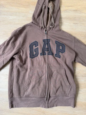 Brun hoodie med dragkedja från GAP - Brun hoodie från GAP med stor logga framtill i svart. Tröjan har dragkedja, två fickor fram och en klassisk huva med snören. Tillverkad i mjukt bomullsmaterial som är perfekt för chill dagar. Enkel och stilren design som funkar till det mesta.
