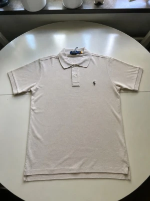 Beige pikétröja från Polo Ralph Lauren - Klassisk beige pikétröja från Polo Ralph Lauren med korta ärmar och krage med två knappar. Tröjan har den ikoniska broderade loggan på bröstet och är tillverkad i mjuk bomull. Perfekt till en avslappnad men stilren look.