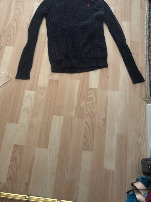 Mörkblå stickad tröja med röd logga - Mörkblå stickad tröja med kabelstickat mönster och lång ärm. Tröjan har ribbade muddar vid ärmslut och nederkant samt en liten röd logga broderad på bröstet. Perfekt för lager på lager och en klassisk look.