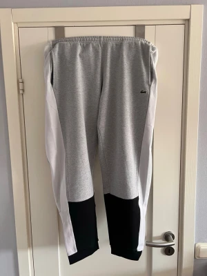 Gråa mjukisbyxor från Lacoste - Snygga grå mjukisbyxor från Lacoste med vita paneler längs sidorna och svarta detaljer vid bensluten. Byxorna har en relaxed fit och elastisk midja för extra komfort. Liten Lacoste-logga på vänster sida. Perfekta för chill dagar.