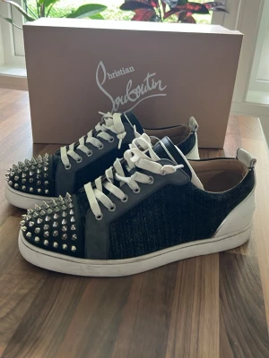 Christian Louboutin svarta sneakers med nitar - Unika svarta sneakers från Christian Louboutin med vita detaljer och vita skosnören. Skorna har en cool design med silverfärgade nitar på tån och en mix av läder och textilmaterial. Perfekta för dig som vill sticka ut med din stil.