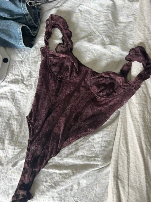 Vinröd spetsbody med breda axelband - Säljer en vinröd body i spets med breda axelband och bygelkupor. Bodyn har ett transparent mönster och markerad midja som ger en snygg siluett. Perfekt att styla under en kavaj eller till jeans. Materialet är stretchigt och följsamt.