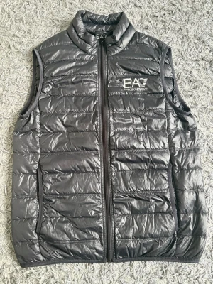 Svart dunväst från EA7 Emporio Armani - Snygg svart dunväst från EA7 Emporio Armani med hög krage och dragkedja framtill. Västen har två sidofickor med dragkedja och ett stilrent EA7-logotryck på bröstet. Lättviktigt material och quiltad design som ger en modern look.