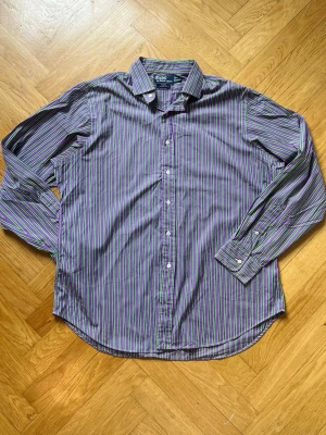 Randig skjorta från Ralph Lauren - Snygg randig skjorta från Ralph Lauren i blått, grönt och lila. Klassisk krage, knäppning framtill och långa ärmar med manschetter. Tillverkad i mjuk bomull som känns skön mot huden. Perfekt för dig som gillar en färgglad och stilren look.