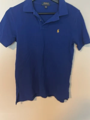 Blå pikétröja från Polo Ralph Lauren - Klassisk blå pikétröja från Polo Ralph Lauren med korta ärmar och krage.