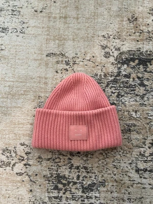 Rosa ribbstickad mössa från Acne Studios - Säljer en snygg rosa ribbstickad mössa från Acne Studios med uppvikt kant och den klassiska Acne-loggan framtill.