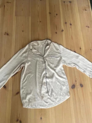 Beige satinpyjamas med svarta detaljer - Tvådelad pyjamas i ljusrosa med svarta detaljer i ett mjukt och lent material.