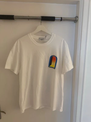Vit Casablanca t-shirt med färgglatt tryck - Säljer en vit t-shirt från Casablanca med ett färgglatt grafiskt tryck i blått, rött, gult och svart på bröstet och ryggen. T-shirten har rund hals och korta ärmar. Perfekt för dig som gillar stilrena plagg med en twist. Storlek L passar M också. 