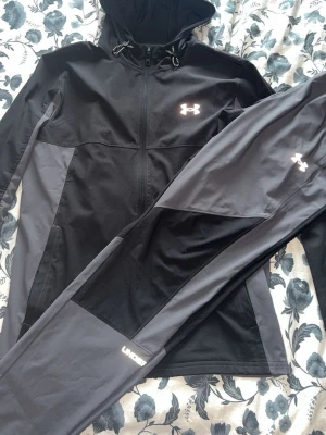 Svart och grå träningsset Under Armour - Snyggt träningsset från Under Armour med svart och grå zip-hoodie och matchande byxor. Setet har dragkedja, huva med snörning och logga på både jacka och byxa. Tillverkat i ett stretchigt funktionsmaterial som passar perfekt till träning eller sport.