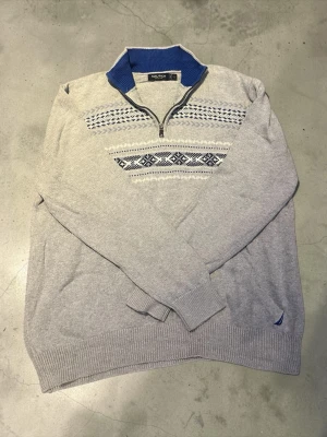 Grå stickad tröja från Nautica - Säljer en grå stickad tröja från Nautica med blått och vitt mönster över bröstet. Tröjan har halv dragkedja, blå ribbad krage och långa ärmar. Skickas inom 24 timmar🚀📦.