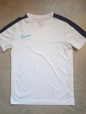 Nike dry fit - Snygg vit sporttröja från Nike med svarta paneler längs axlarna och ett ljusblått Nike-märke på bröstet. Tillverkad i ett lätt och ventilerande funktionsmaterial som passar perfekt till träning eller match.