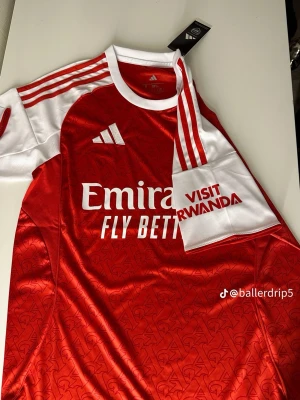 Arsenal matchtröja röd/vit Adidas - Snygg Arsenal matchtröja från Adidas i rött och vitt med klubbmärke, Adidas-logga och sponsortryck. Tröjan har vita detaljer på axlar och ärmar samt ett subtilt mönster i tyget. Perfektp för fotbollsfans och träning.