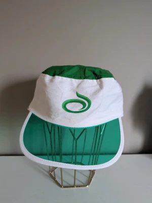 Grön och vit retrokeps med plastskärm - Säljer en unik retrokeps i vitt och grönt med en genomskinlig grön plastskärm. Kepsen har ett grönt tygparti upptill och ett tryckt grönt märke framtill. Perfekt för dig som gillar vintage och vill sticka ut med en cool accessoar.