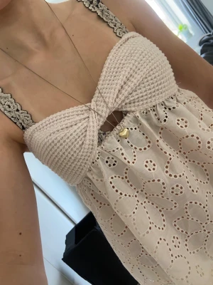 Beige mönstrad tube top med virkad byst - Supersöt beige linnetopp med virkad byst och breda spetsband. Toppen har broderade blommor och hålmönster i tyget, samt en twistad detalj framtill. Perfekt för varma dagar och ger en romantisk vibe.