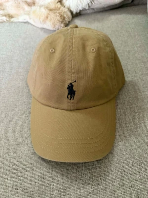 Beige keps från Polo Ralph Lauren - Snygg beige keps från Polo Ralph Lauren med klassisk böjd skärm och justerbart spänne bak. Framtill finns den ikoniska broderade polospelaren i mörkblått och baktill står det 'POLO' broderat. Tillverkad i bomull för en skön och avslappnad look.