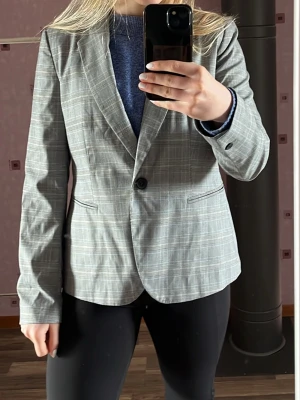 Grå rutig kavaj med enkelknäppning - Stilren grå kavaj med diskret rutmönster och enkelknäppning. Klassisk passform med två fickor framtill och smala slag. Perfekt till både jeans och kostymbyxor. Materialet känns lätt och smidigt, vilket gör den enkel att bära hela dagen. Kavajen är mycket liten i storlek men skulle motsvaras med en M