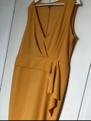 Gul omlottklänning med volang - Snygg gul omlottklänning med v-ringning och volangdetalj i midjan. Klänningen är ärmlös och har en elegant, figurnära passform som ger en modern siluett. Perfekt för dig som vill sticka ut med färg och stil.