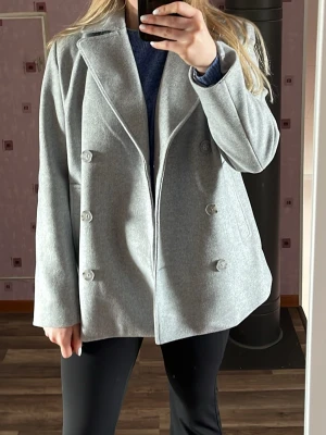 Ljusgrå dubbelknäppt kappa - Snygg ljusgrå rock med dubbelknäppning och breda slag. Rocken har en rak passform och är tillverkad i ett mjukt ullblandat material. Perfekt för kyliga dagar och enkel att matcha med olika outfits.