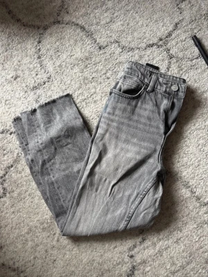 Grå raka jeans med rå kant - Säljer ett par grå jeans med rak passform och rå, fransig kant nedtill. Jeansen har klassisk femficksdesign och är tillverkade i denim. Perfekta för en avslappnad och trendig look.